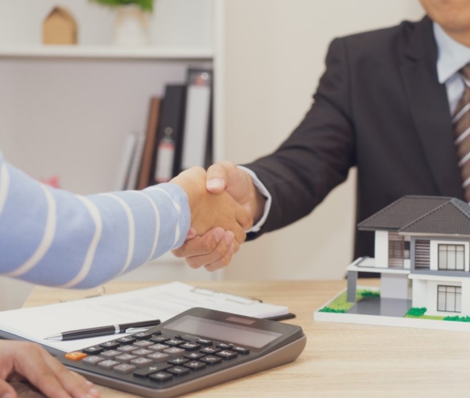 Cierre de acuerdo inmobiliario entre comprador y asesor, con apretón de manos, calculadora, documentos y maqueta de casa sobre la mesa
