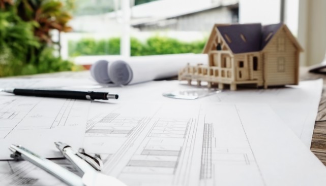 Planos arquitectónicos sobre una mesa con herramientas de dibujo técnico y una maqueta de casa en miniatura